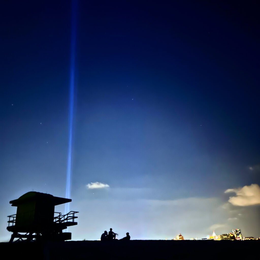 An image of a silhouette  of a lifeguard tower at sunset. Shooting up from the base of the tower are two beams of light. There is dark navy blue sky with the pale yellow glow of a city in the far distance a group of people sitting in the center. 