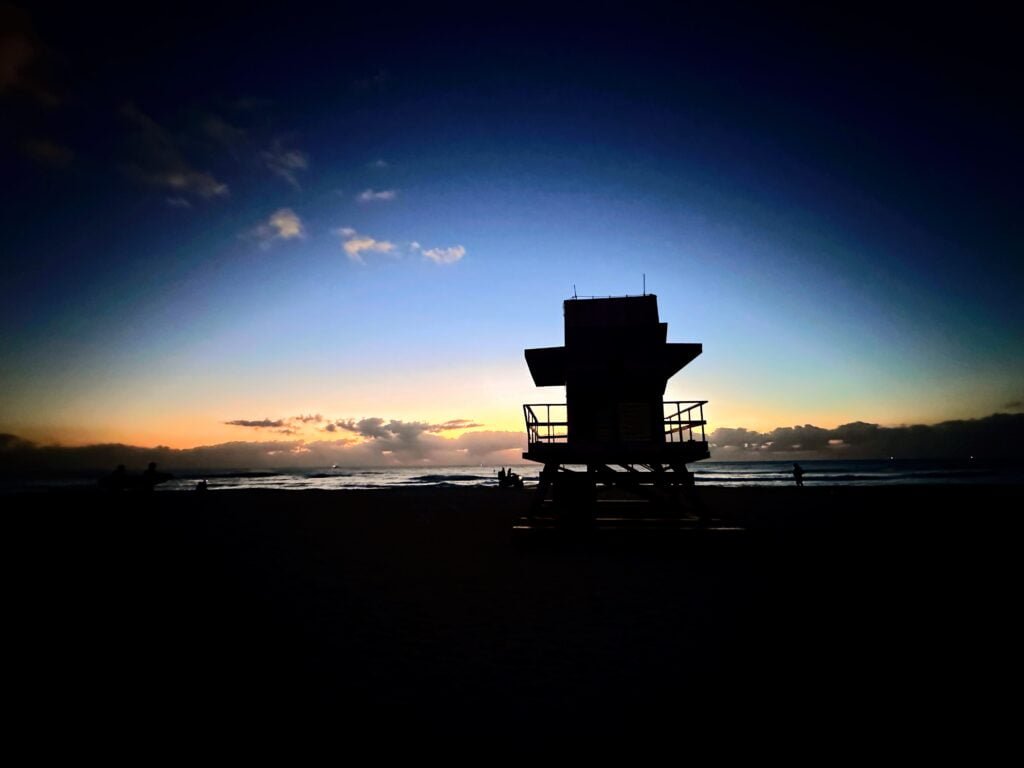 An image of a sunrise and the backside of lifeguard tower on the beach, facing the ocean. In the distance,  there is a deep navy blue sky fading downward into an amber glow on the horizon. 